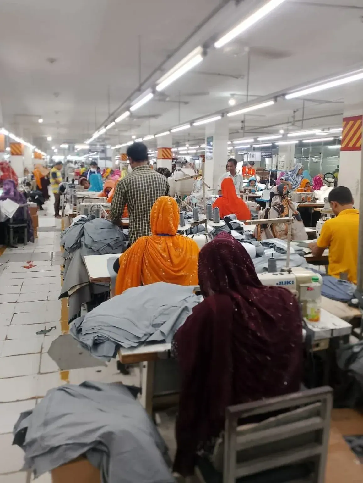 Apparel Sewing Line with Fabric Panels Sewing operators working beside stacks of grey fabric panels on a production line.