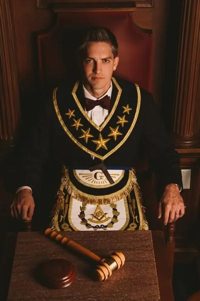 Masonic Regalia Collar and Apron Reference | ZESTLET A person wearing a black and gold ceremonial collar and embroidered apron beside a gavel.